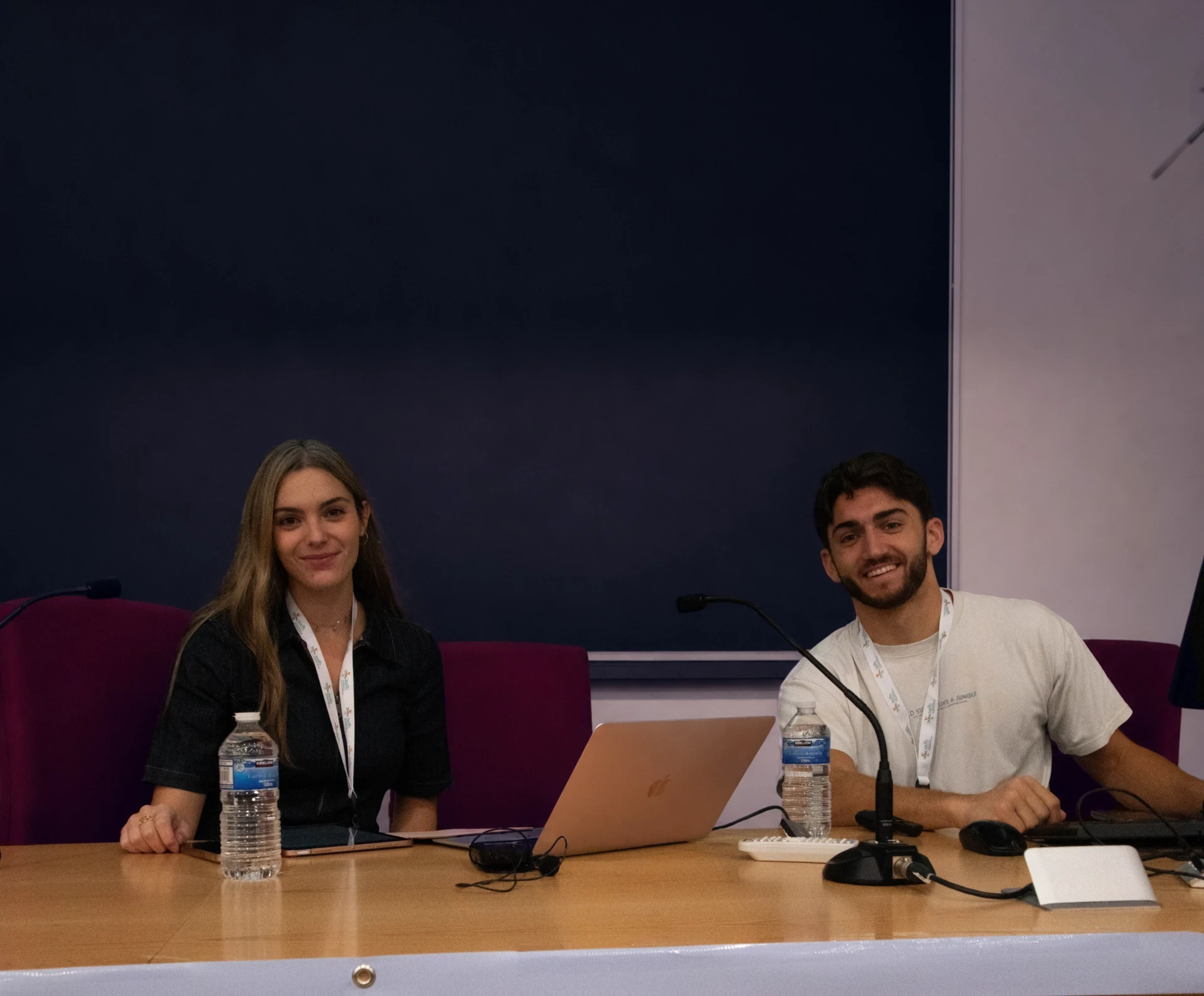 Two people sitting at a speakers table with microphones and an open laptop, smiling at the camera during a training day