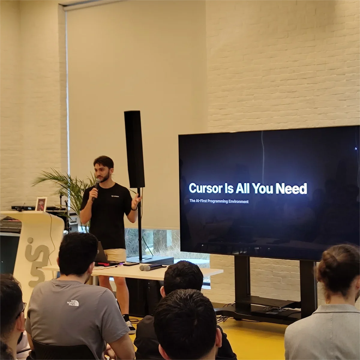 Un ponente con micrófono junto a una pantalla que dice «Cursor Is All You Need», ante una sala llena