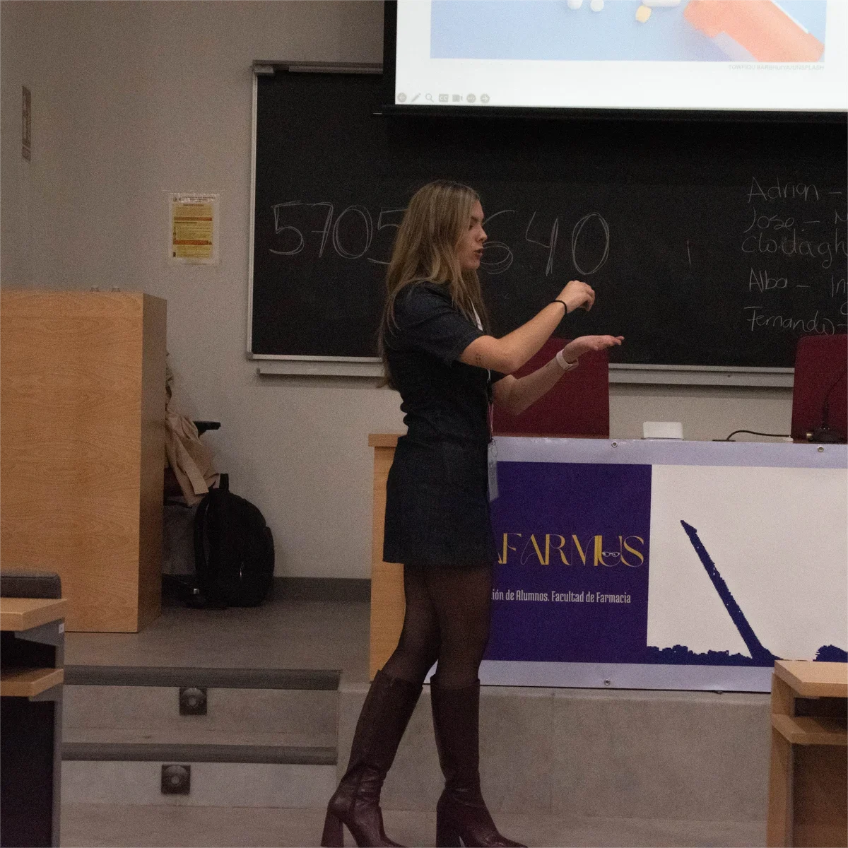 Clase en un aula universitaria: una profesora explicando junto a una pizarra con cifras escritas a mano