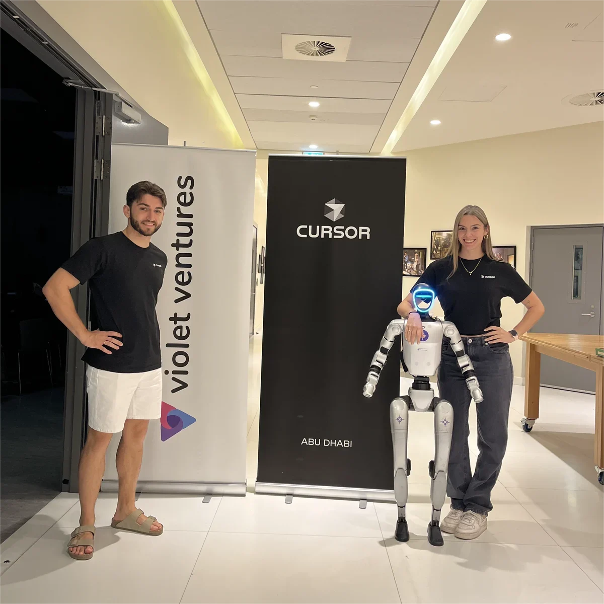 Dos integrantes del equipo junto a un robot humanoide, delante de los carteles de Cursor en Abu Dhabi