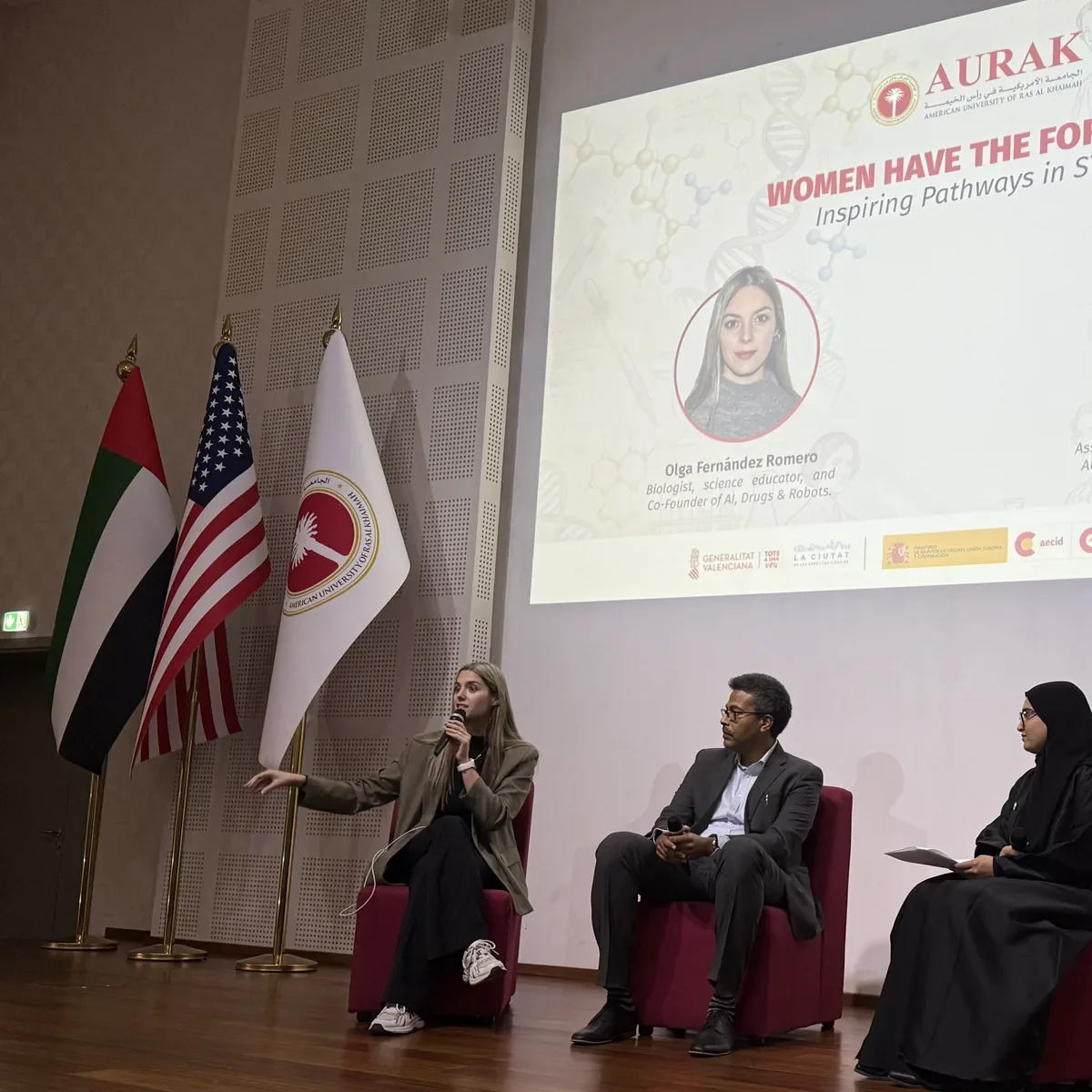 Una ponente con micrófono en una mesa redonda de la universidad AURAK, en Emiratos, bajo una pantalla con su nombre