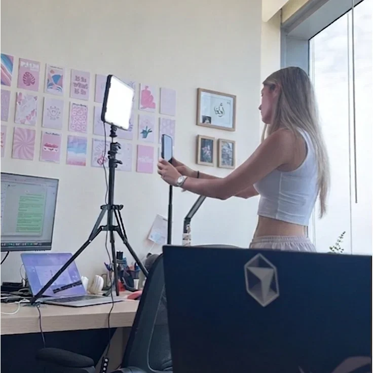 Una integrante del equipo grabando con el móvil sobre un trípode, con anillo de luz y dos pantallas encendidas en el escritorio